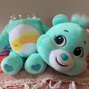 May include: A teal plush bear with a glass jar head. The bear has a white belly with a yellow and orange rainbow and a smiling star. The bear has a blue heart on its foot and a blue heart-shaped nose.