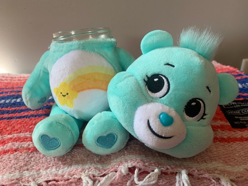 May include: A teal plush bear with a glass jar head. The bear has a white belly with a yellow and orange rainbow and a smiling star. The bear has a blue heart on its foot and a blue heart-shaped nose.