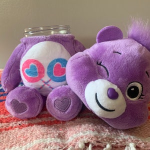 May include: A purple plush bear with a white belly featuring pink and blue hearts. The bear is designed to hold a glass jar. The bear has a smiling face and a heart-shaped detail on its foot. A jar is inserted into the bear's body.