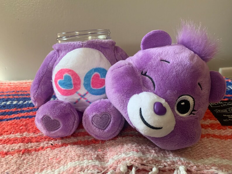 May include: A purple plush bear with a white belly featuring pink and blue hearts. The bear is designed to hold a glass jar. The bear has a smiling face and a heart-shaped detail on its foot. A jar is inserted into the bear's body.