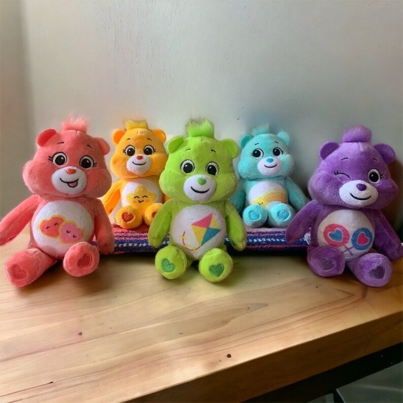 May include: Five colorful Care Bear plush toys are arranged on a wooden surface. The bears are pink, yellow, green, blue, and purple. Each bear has a unique belly badge and a smiling face. They are sitting on a striped mat.