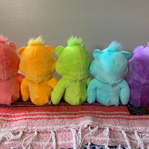 May include: Five colorful plush bear toys in a row, each a different shade: pink, orange, yellow, blue, and purple. The bears are sitting on a striped blanket with fringe.
