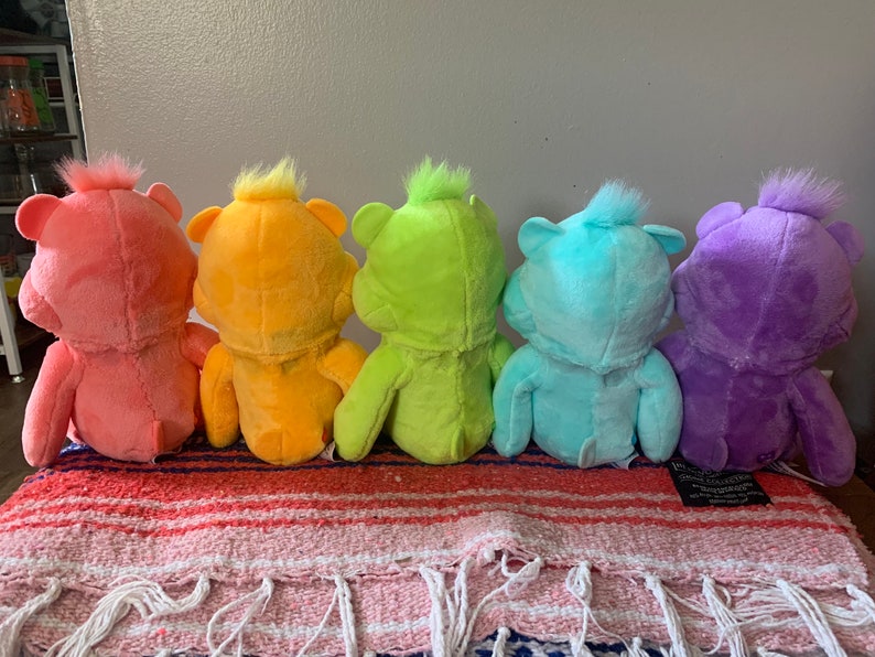 May include: Five colorful plush bear toys in a row, each a different shade: pink, orange, yellow, blue, and purple. The bears are sitting on a striped blanket with fringe.
