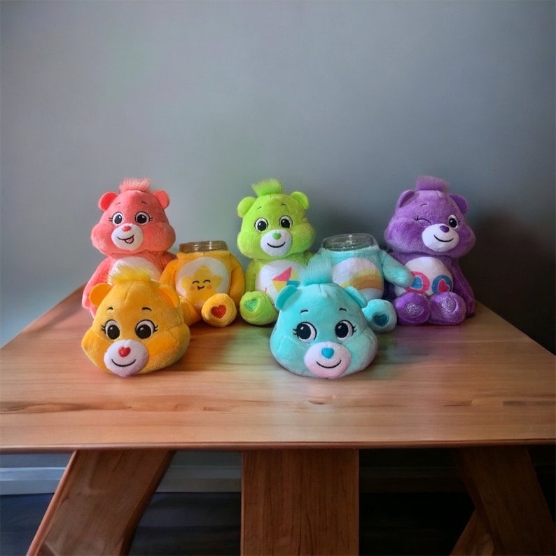 May include: A collection of colorful plush Care Bears, including pink, yellow, green, blue, and purple. Each bear has a unique design and expression. Some bears have glass jars as bodies, while others are complete plush figures. They are arranged on a wooden table.