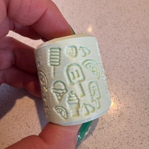 May include: A pale green cylindrical object featuring embossed summer designs, such as ice cream cones, ice lollies, and fruit slices. The object is held by a person.