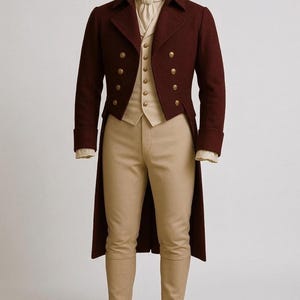 May include: A burgundy double-breasted coat with gold buttons, paired with a cream-colored vest, a white cravat, and tan trousers. The coat has a long, tailored silhouette, suggesting formal attire. The outfit is displayed on a mannequin.