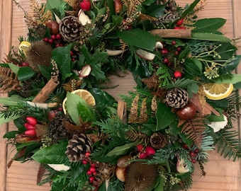 May include: A circular wreath composed of various natural elements. It features green leaves, pine cones, red berries, dried orange slices, cinnamon sticks, and other dried foliage. The wreath is arranged on a wooden surface.