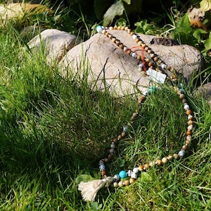 Puede incluir: Un collar de cuentas marrones y blancas con una cuenta turquesa y un borla cuelga de una roca en una zona cubierta de hierba.