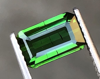 May include: A vibrant green gemstone with a rectangular cut, held in tweezers. The gemstone has a slight golden hue.