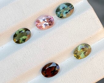 May include: Five oval-shaped gemstones in various shades of green, brown, and pink. The gemstones are arranged in a row on a white surface.