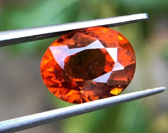 May include: A close-up of an orange gemstone held by tweezers. The gemstone is oval-shaped and has a faceted cut.