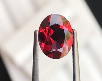 May include: A deep red oval gemstone held by tweezers against a white background.