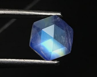 May include: A blue iridescent gemstone with a faceted, hexagonal shape. The gemstone is held by tweezers against a black background.