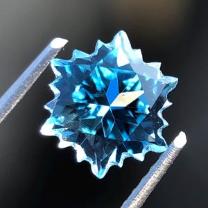 Könnte beinhalten: Ein sternförmiger, facettierter Edelstein in leuchtendem Blau, gehalten von einer Pinzette. Die Facetten des Steins reflektieren das Licht und erzeugen ein funkelndes Aussehen. Die scharfen Kanten des Edelsteins heben sich vor dem dunklen Hintergrund ab.