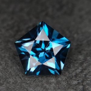 May include: A faceted gemstone in a pentagonal shape, featuring a star-shaped design within. The gem has a deep teal color with lighter, reflective facets. The gemstone is set against a dark gray background.