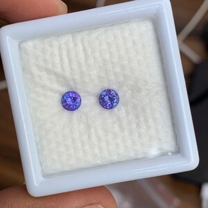K&ouml;nnte beinhalten: Zwei runde, violette Edelsteine ​​werden in einer wei&szlig;en, mit Samt ausgekleideten Schachtel pr&auml;sentiert. Die Edelsteine ​​sind in runder Form gefasst und haben eine glatte, polierte Oberfl&auml;che.