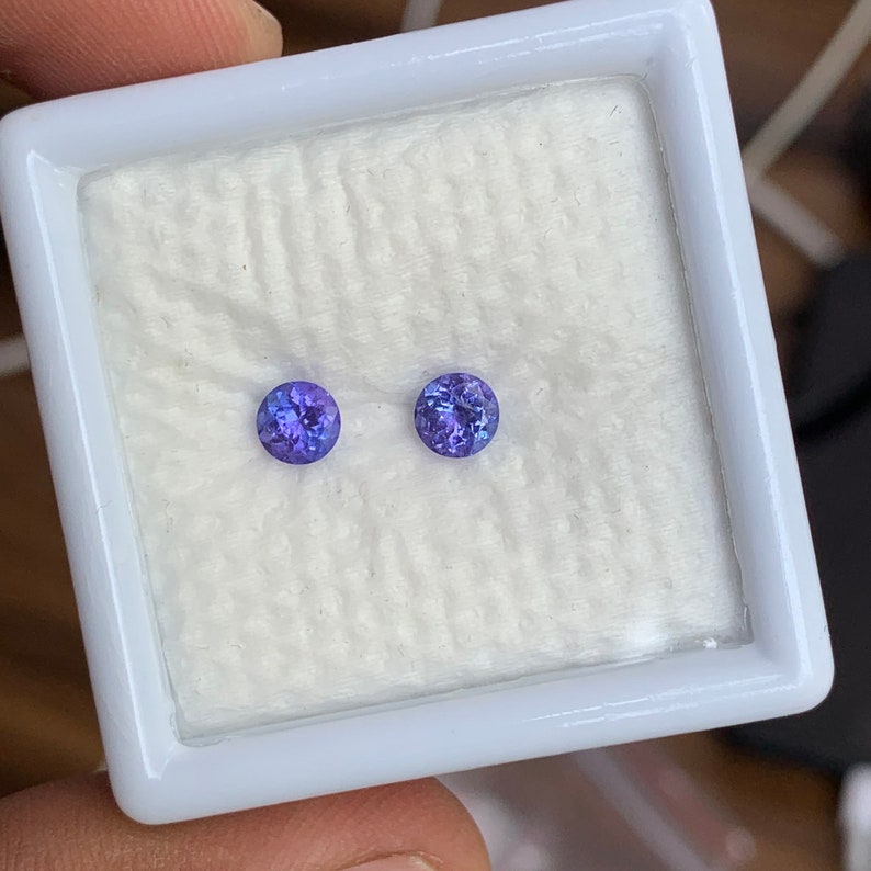 K&ouml;nnte beinhalten: Zwei runde, violette Edelsteine ​​werden in einer wei&szlig;en, mit Samt ausgekleideten Schachtel pr&auml;sentiert. Die Edelsteine ​​sind in runder Form gefasst und haben eine glatte, polierte Oberfl&auml;che.