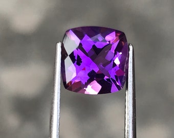 Könnte beinhalten: Ein facettierter, quadratisch geschliffener Amethyst-Edelstein, der von einer Metallpinzette gehalten wird. Der Edelstein hat eine tiefviolette Farbe mit mehreren Facetten, die das Licht reflektieren. Der Hintergrund ist ein verschwommenes Grau.
