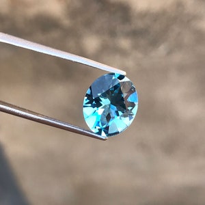 May include: A light blue oval-shaped gemstone held by tweezers against a blurred background.