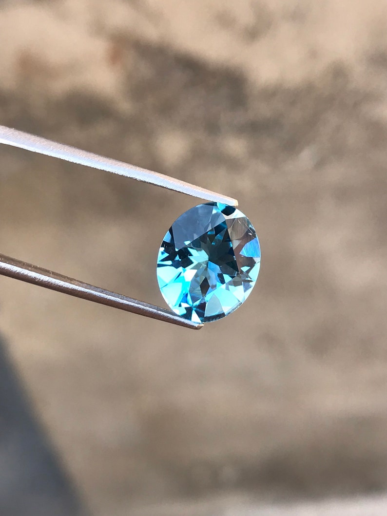 May include: A light blue oval-shaped gemstone held by tweezers against a blurred background.
