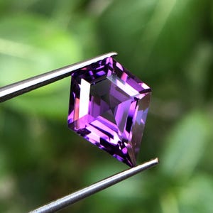 Peut inclure: Une pierre précieuse d'améthyste violette foncée avec une taille carrée et une finition facetée. La pierre précieuse est tenue dans des pinces sur un fond vert.