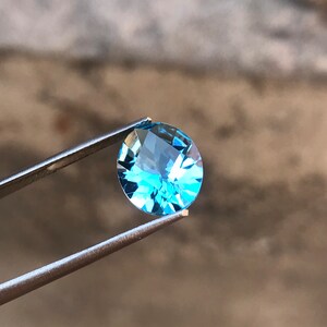 May include: A light blue oval-shaped gemstone held by tweezers against a blurred background.