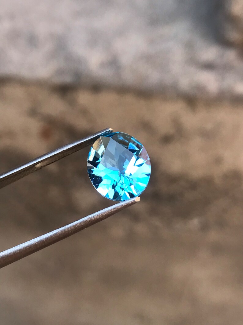 May include: A light blue oval-shaped gemstone held by tweezers against a blurred background.