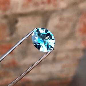 May include: A light blue oval-shaped gemstone held by tweezers against a blurred brick background.