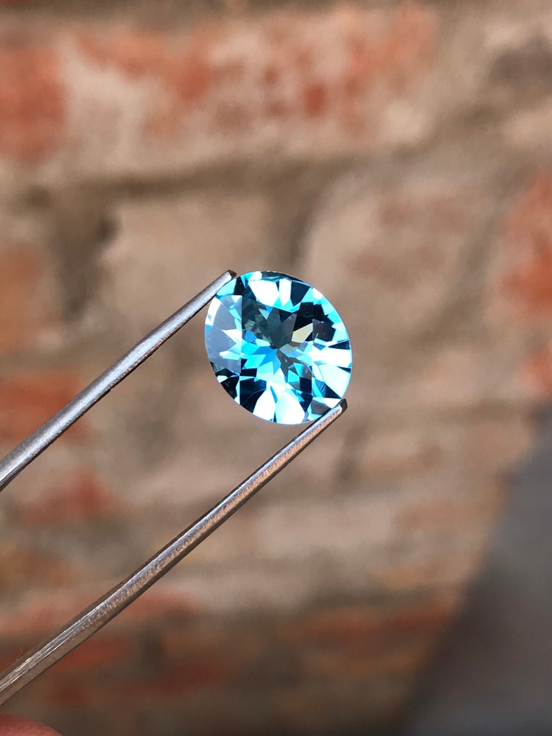 May include: A light blue oval-shaped gemstone held by tweezers against a blurred brick background.