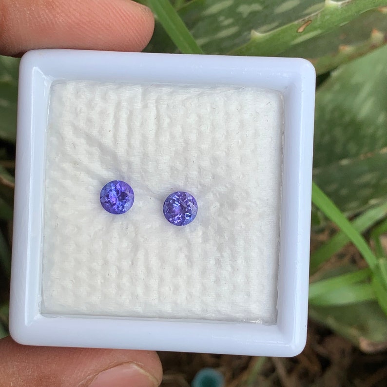 K&ouml;nnte beinhalten: Zwei runde, violette Edelsteine in einer wei&szlig;en, quadratischen, samtenen Box.