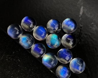 Op de afbeelding: Een groep van 12 ronde, iriserende maansteen cabochons in verschillende tinten blauw en wit. De stenen zijn in een driehoekige vorm op een zwart oppervlak gerangschikt.