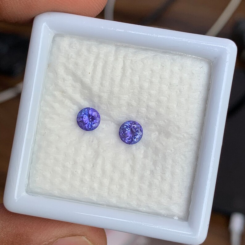 K&ouml;nnte beinhalten: Zwei runde, violette Edelsteine in einer wei&szlig;en, quadratischen, mit Samt ausgekleideten Schachtel.