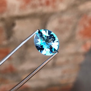May include: A light blue oval-shaped gemstone with facets, held by tweezers against a blurred background.
