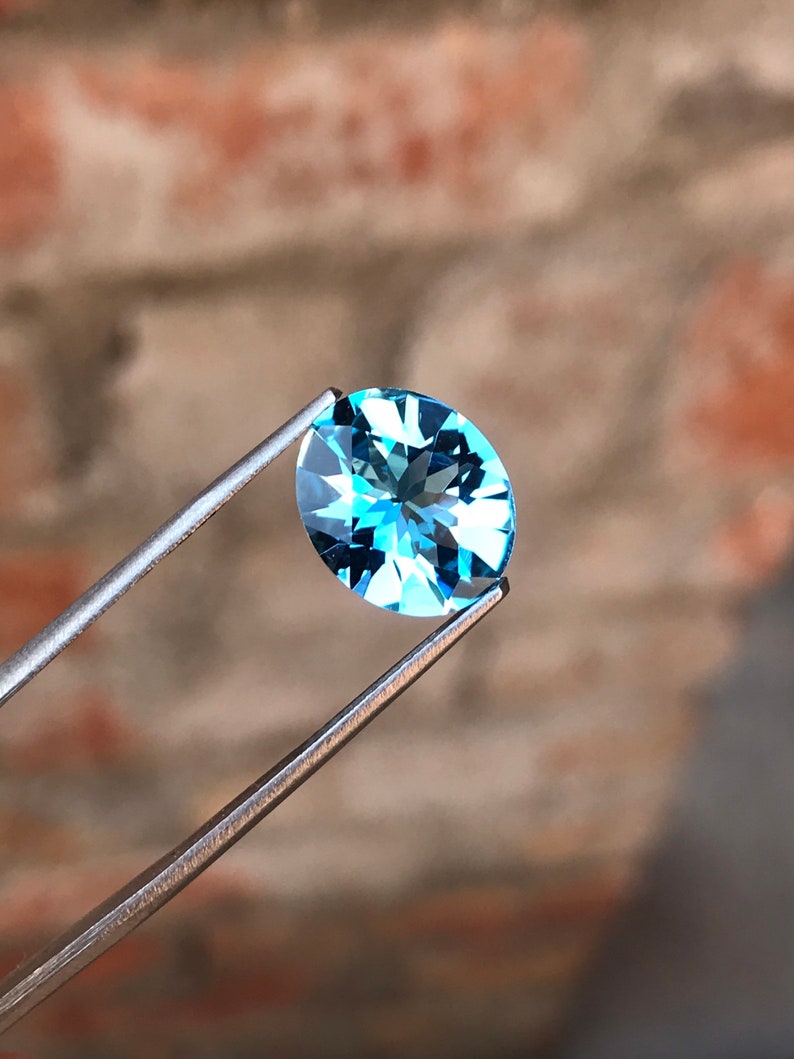 May include: A light blue oval-shaped gemstone with facets, held by tweezers against a blurred background.