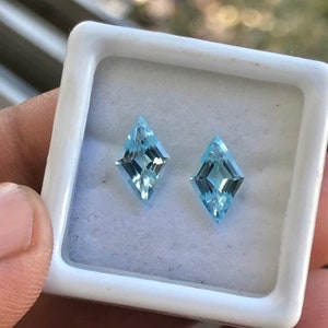 May include: Two light blue, kite-shaped gemstones in a white box. The gemstones are faceted and have a clear, glassy appearance.