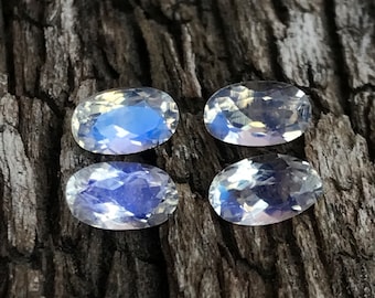 May include: Four oval-cut moonstone gemstones with a blue and white iridescent glow. The stones are set against a dark, textured wooden surface, highlighting their natural beauty. These gemstones are ideal for jewelry making.