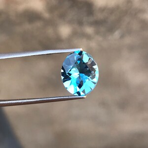 May include: A light blue oval-shaped gemstone held by tweezers against a blurred background.