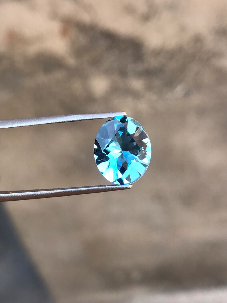 May include: A light blue oval-shaped gemstone held by tweezers against a blurred background.