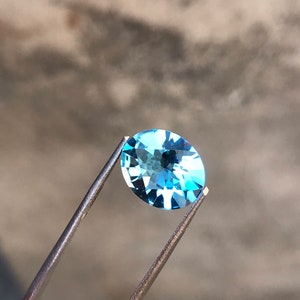 May include: A light blue oval-shaped gemstone held by tweezers against a grey background.