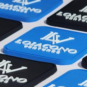 Peut inclure: Plusieurs dessous de verre carrés, bleus et noirs. Chaque dessous de verre porte l'inscription blanche "LOJACONO VENTURES" et un logo "LV" stylisé. Les dessous de verre sont disposés sur une surface blanche.