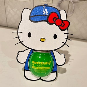 以下が含まれることがあります： 青いキラキラ光る野球帽と赤いリボンが付いた、白いキラキラ光るハローキティ型のワインクーラーホルダー。 ホルダーには、緑色のBuzzBallz Lime 'Rita Chillerが入っています。 クーラーのテキストは「LIME 'RITA CHILLER」と表示されています。