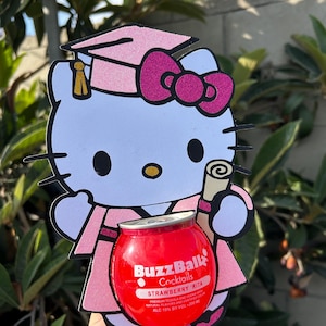 Może przedstawiać: Różowa i biała dekoracja na ukończenie szkoły w kształcie Hello Kitty trzymająca czerwony koktajl BuzzBallz Strawberry Rita. Dekoracja przedstawia czapkę i szatę absolwenta.