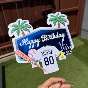 May include: A cake topper featuring a blue holographic sign with the text Happy Birthday, two green palm trees, a baseball, a wooden bat, and a white jersey with the name JESSE and the number 80, alongside a blue LA logo emblem.