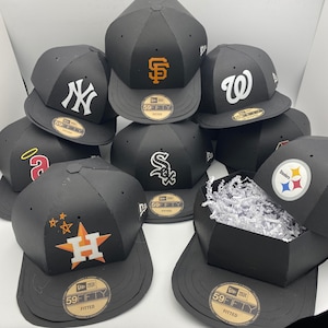 May include: A collection of black baseball caps with various team logos, including the New York Yankees, San Francisco Giants, and Houston Astros. The caps are stacked, with one open box filled with white paper. Each cap features the "59FIFTY" and "FITTED" labels.