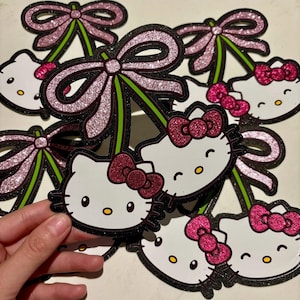 Puede incluir: Varias decoraciones con forma de cereza con el tema de Hello Kitty. Cada decoración presenta una cara blanca de Hello Kitty con un lazo rosa, ojos amarillos y un contorno negro. Las cerezas tienen lazos de purpurina rosa y tallos verdes.