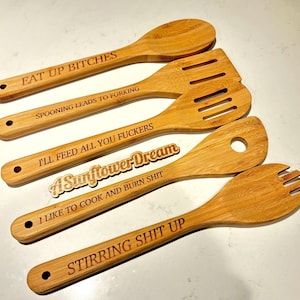 May include: A set of five wooden kitchen utensils with humorous phrases engraved on them. The utensils include a spoon, slotted spatula, and a fork-like tool. The text includes "EAT UP BITCHES" and "STIRRING SHIT UP".