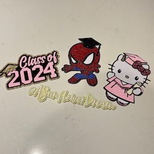 以下が含まれることがあります： 卒業キャップをかぶったスパイダーマン、卒業ガウンを着たハローキティ、そして「Class of 2024」の文字が特徴のキラキラした切り抜き。筆記体で「@ASunflowerDream」と書かれています。