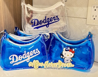 Op de afbeelding: Transparante en blauwe vinyl schoudertassen met witte ritsen. De tassen tonen het woord "Dodgers" in witte letters. Eén tas heeft een Hello Kitty-afbeelding met een Dodgers honkbalpet. De tassen staan op een wit oppervlak.