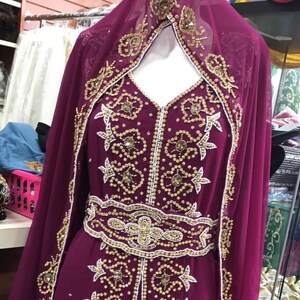 May include: A long, burgundy dress with gold and white beading. The dress has a high neckline and long sleeves. The dress is decorated with intricate floral patterns.