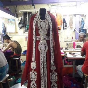 May include: A burgundy-colored, floor-length gown with intricate silver and gold embroidery. The dress features a high neckline and long sleeves, with a matching shawl. The garment is displayed on a mannequin in a tailor shop.